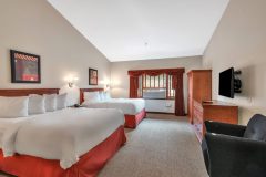 Two queen beds with white linens and red pillows and red blanket in front of sofa chair and television at The Inn at Ohio Northern University in Ada, OH