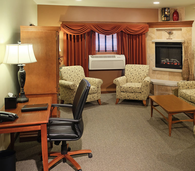 Group of white armchairs near air conditioner near wooden coffee table in front of wooden dresser and wooden desk with black leather chair and fireplace at The Inn at Ohio Northern University in Ada, OH