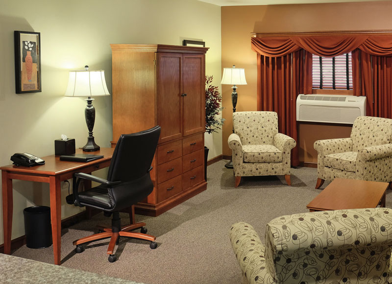 Group of white armchairs near air conditioner near wooden coffee table in front of wooden dresser and wooden desk with black leather chair at The Inn at Ohio Northern University in Ada, OH