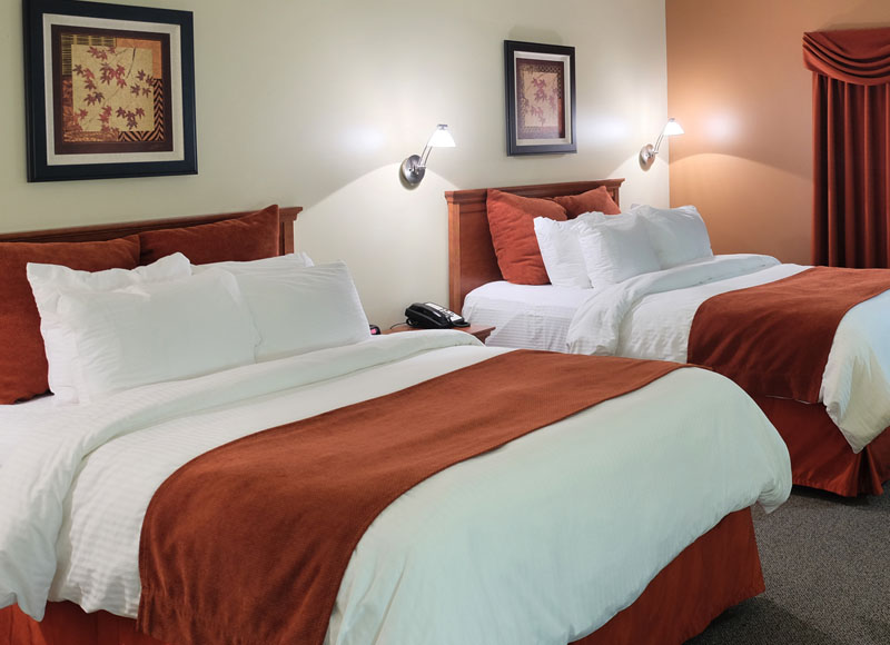 Two queen beds with white linens and red pillows and red blanket near side table and art at The Inn at Ohio Northern University in Ada, OH