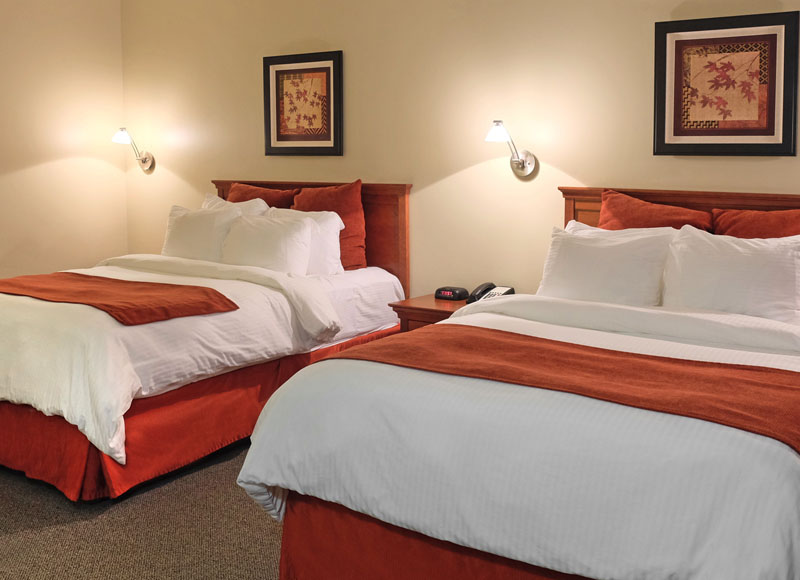 Two queen beds with white linens and red pillows and red blanket near side table and art at The Inn at Ohio Northern University in Ada, OH