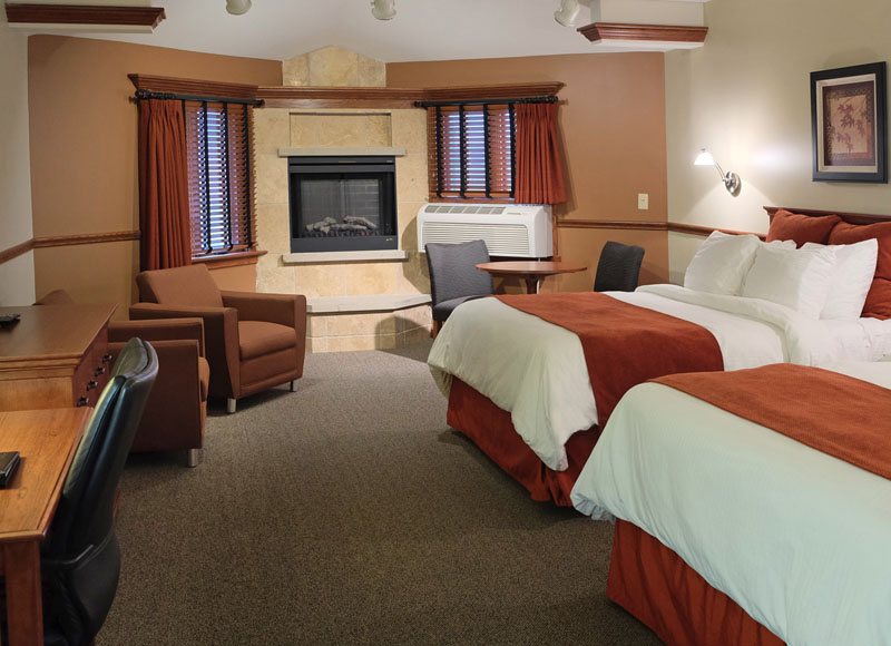 Large bed with white linens and red blanket in front of desk with black leather chair near stone fireplace and brown armchairs at The Inn at Ohio Northern University in Ada, OH