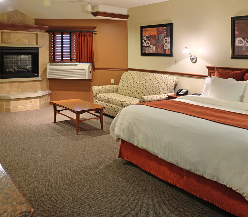 Large bed with white linens and red blanket near coffee table in front of white couch and large air conditioner near stone fireplace at The Inn at Ohio Northern University in Ada, OH