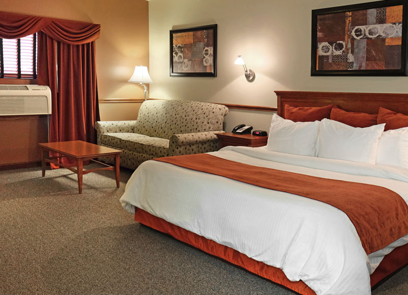 Large bed with white linens and red blanket near coffee table in front of white couch and large air conditioner at The Inn at Ohio Northern University in Ada, OH