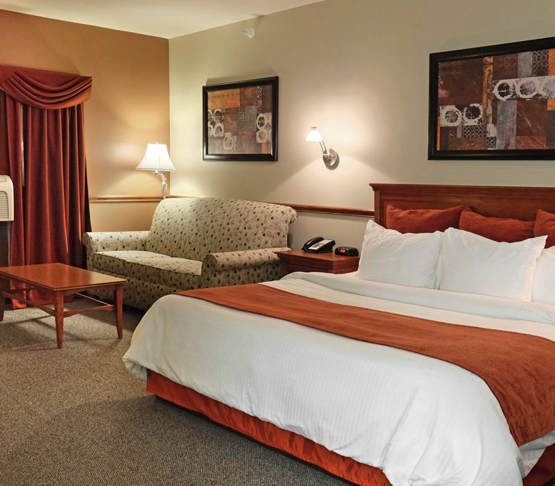 Large bed with white linens and red blanket near coffee table in front of white couch and large air conditioner at The Inn at Ohio Northern University in Ada, OH