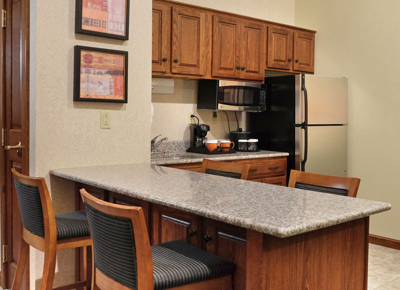 Granite counter top with bar stools near refrigerator and microwave with coffeemaker and wooden cabinets at The Inn at Ohio Northern University in Ada, OH