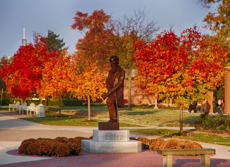 Statue of man in front of fall foliage and stone pathway with stone bench in front and green lawn at The Inn at Ohio Northern University in Ada, OH