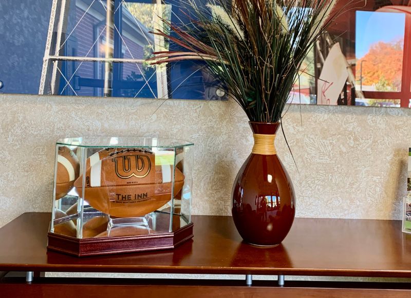 Wooden table with vase on top containing plant in front of painting next to Wilson football in glass case in The Inn at Ohio Northern University in Ada, OH