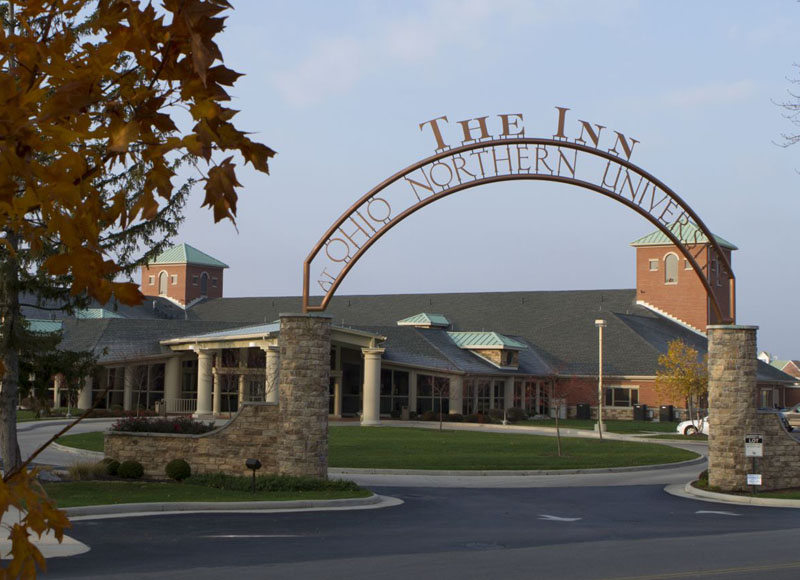 Outdoor Entrance to The Inn at Ohio Northern University in Ada, OH with sign hanging above stone wall and driveway in front of brick building near trees